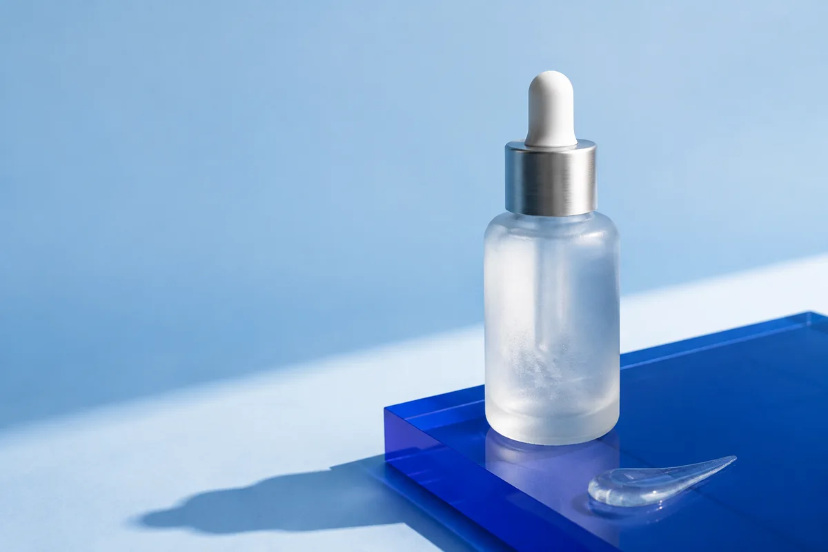 An illustrative skincare campaign: a frosted serum bottle on blue acrylic in sunlight